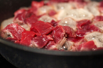 Chunks of raw beef are sizzling in a dark pan, with steam and bubbles rising from the meat as it cooks