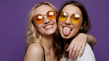 Trendy young women with retro sunglasses embracing and making funny faces against a purple background