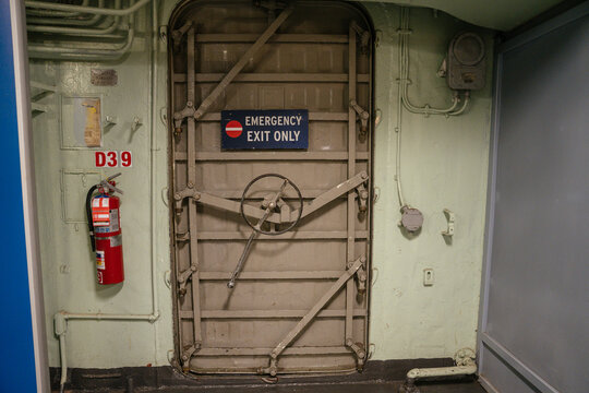 A secure emergency exit door with a wheel-lock mechanism is mounted on a ship or industrial wall. A red fire extinguisher is attached beside the hatch labeled.