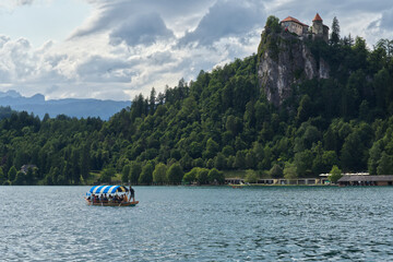 A picturesque scene at Lake Bled, Slovenia, featuring a traditional Pletna boat on the serene blue water with Bled Castle perched majestically atop a lush, forested cliff under a dramatic sky.