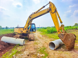 A heavy yellow excavator is digging near large concrete pipes in a field