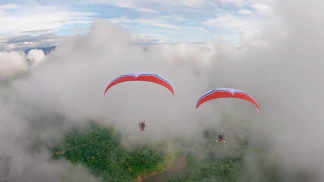 Two paragliders soar through the clouds over a mountainous landscape