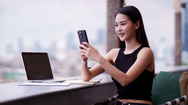 Asian adult young businesswoman uses smartphone laptop for remote work at modern urban cafe enjoying city view