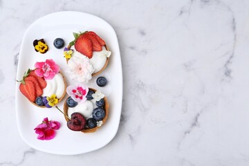 Sweet tartlets with berries and flowers on light marble table, top view with space for text. Delicious dessert