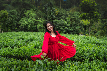 A woman in a red dress and sunhat enjoys a peaceful moment in a lush tea garden surrounded by green hills and nature.

