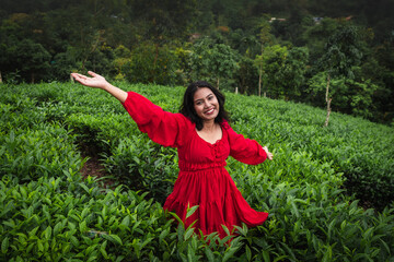 A woman in a red dress and wide-brimmed hat enjoys a peaceful moment in a lush green tea garden surrounded by misty hills and nature.

