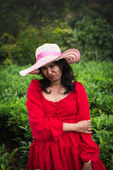 A woman in a red dress and wide-brimmed hat enjoys a peaceful moment in a lush green tea garden surrounded by misty hills and nature.

