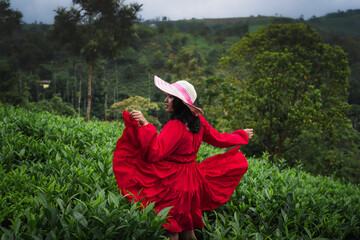 A woman in a red dress and wide-brimmed hat enjoys a peaceful moment in a lush green tea garden surrounded by misty hills and nature.

