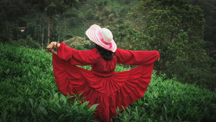 A woman in a red dress and wide-brimmed hat enjoys a peaceful moment in a lush green tea garden surrounded by misty hills and nature.

