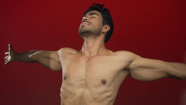 Man with bare chest and outstretched arms in a red studio setting showing muscular torso and shoulders; serenity.
