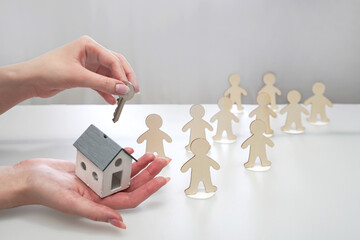 Hand holding a model of a house and a key, offering it to figures of people in a line. Housing distribution, access to home ownership, mortgage, renting a house