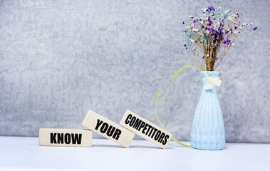 KNOW YOUR COMPETITORS text on wooden blocks on a gray background next to a flower