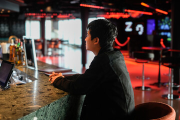 A person sits contemplatively at a bar counter with vibrant lighting and glass decor, conveying a serene and stylish atmosphere in a modern bar setting