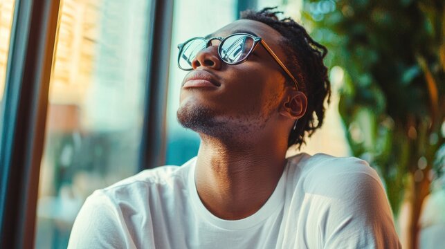 A young man with dreadlocks, wearing a white t-shirt, sitting in a cafe with a laptop and a cup of coffee, looking out the window