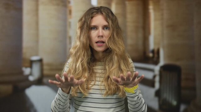 Woman raises hands calming in corridor featuring blonde young subject showing deescalation of tension in portrait indoors.
