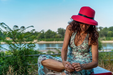 middle-aged woman wearing a red hat sitting on a table removing a thorn from her foot at sunset by...