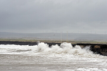 Powerful waves crash against the sea wall with wind turbines for energy production on the horizon