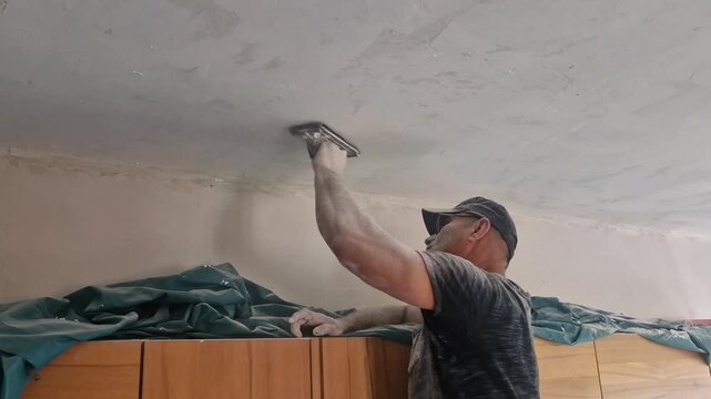 A strong, skilled man sands a repaired ceiling using circular hand motions above his head. Fine plaster dust falls continuously, covering his face and body in white