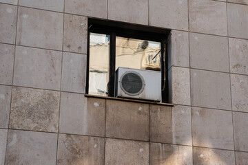 An air conditioning unit is installed in a window of a beige stone building. The unit reflects the...