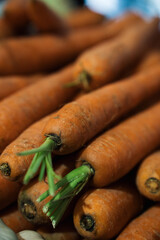 Bright, fresh carrots are stacked at a market, showcasing their vibrant orange color and leafy greens. Shoppers browse the stalls in a lively atmosphere during the day.