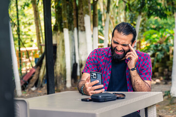 Stressed Colombian man in plaid shirt holding phone and touching head in tropical garden during...