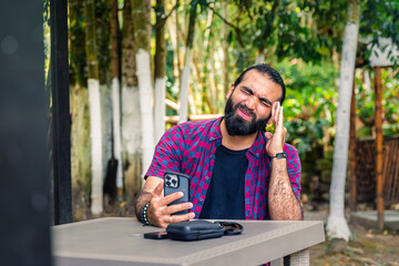 Stressed Colombian man in plaid shirt holding phone and pressing hand to forehead during remote...