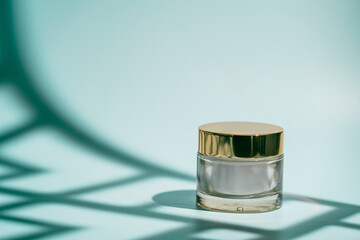 Glass skincare cream jar with golden lid placed on a pastel aqua background with soft light and...