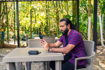Colombian man in plaid shirt using phone while sitting at outdoor table in lush tropical garden in...