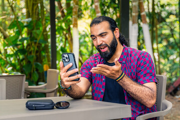 Expressive Colombian man in plaid shirt reacting with dissatisfaction while holding phone at...