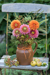 Blumenstrauß mit Dahlien, Gräsern, Wilder Möhre und Himbeerzweigen im rustikalen Krug auf einem Gartenstuhl