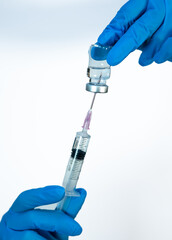 Gloved hands holding a syringe and inverted vial while drawing medication with a needle, shown...