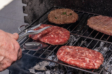 raw hamburger patties on a charcoal grill, just starting to cook. A hand uses metal tongs to position the meat over hot embers and grey ash