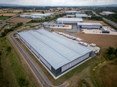 Aerial View of Large Distribution Warehouse and Fleet of Delivery Trucks in Active UK Logistics Park – Modern Freight Hub with Road Access, Surrounded by Green Fields and Industrial Units