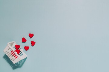 Red heart shapes spill playfully from a white mini shopping basket on soft blue background. Perfect for themes of love, gifting, and caring.
