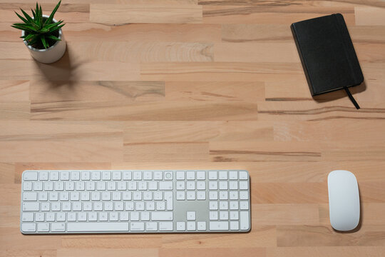 home office workspace of modern wireless white keyboard, wireless white mouse, black notebook on wooden table with a green plant in flowerpot. top view with copy space.