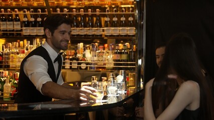 Smart bartender serving gin fizz or tonic with sliced lemon at luxurious bar, engaging lively conversation with VIP customer couple at side view in atmosphere at lighting warm nightclub. Vinosity.