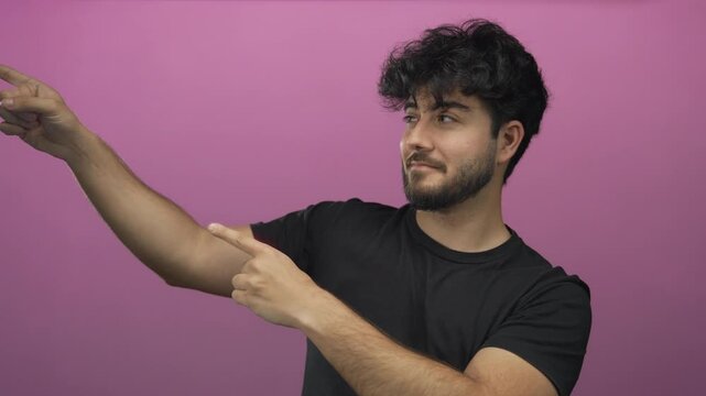 Young man with beard pointing against a vibrant pink background, conveying direction and focus with a confident expression and casual black t-shirt.