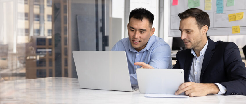 Horizontal photography of two focused, multiracial business companions using tablet device and laptop, negotiates on contract conditions, engaged in task completion or deal discussion in modern office