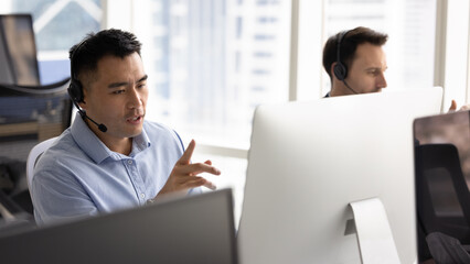 Focused Asian call center or helpline employee looks at PC monitor screen, leads clients...