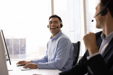 Asian man, call center employee laughing seated at desk with colleague, wearing headset lead...