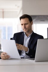 Focused business man learning points of contract, carefully reading, analyzing important details and responsibilities outlined in formal agreement seated at table with open laptop in modern workplace