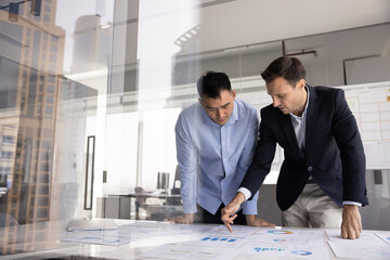 Two multiethnic businessmen standing at meeting desk over heap of papers, prepare report, analyze...