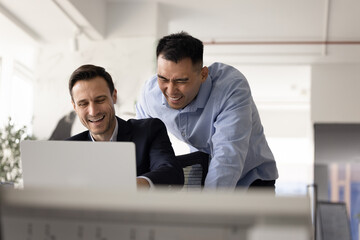 Male teammates watching online content and laughing during break in workplace using laptop, enjoy...