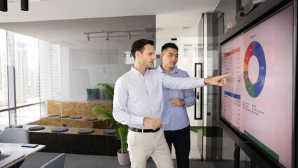 To male teammates engaged in formal business meeting, presentation, or strategy planning in modern conference room, reviewing data displayed on large digital screen with charts and graph. Strategizing