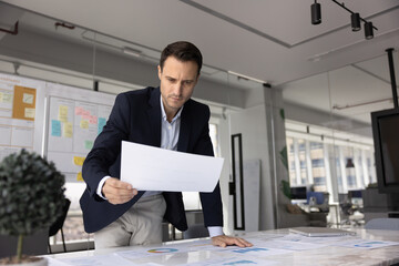 Focused Hispanic business professional stands in modern conference room, examine printed sheet with...