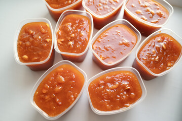 Fresh homemade apricot preserve in multiple plastic containers placed on a white surface in soft...