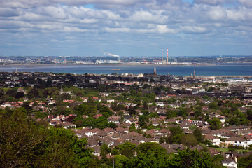 Expansive aerial view of a vibrant Dublin city with diverse rooftops, lush greenery, and a wide bay extending to distant hills under a bright sky