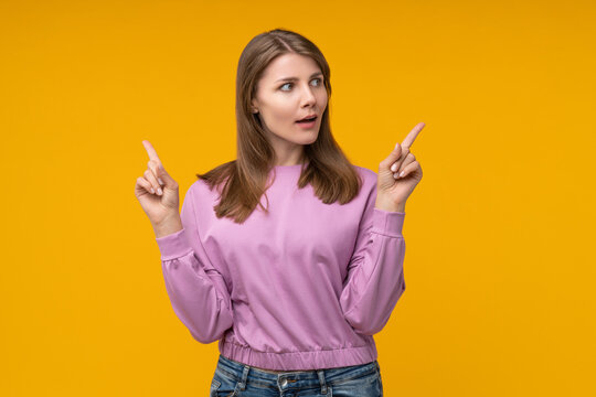 Image of a young woman posing over a yellow studio background pointing upward with both index fingers and looking aside