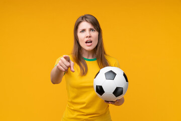 Confident young female soccer fan or player holding a soccer ball and pointing at the camera with an accusing expression on yellow background