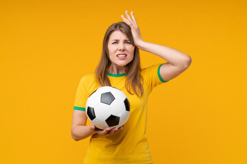 Disappointed young woman football fan in yellow t-shirt holding soccer ball in one hand and touching her head with the other